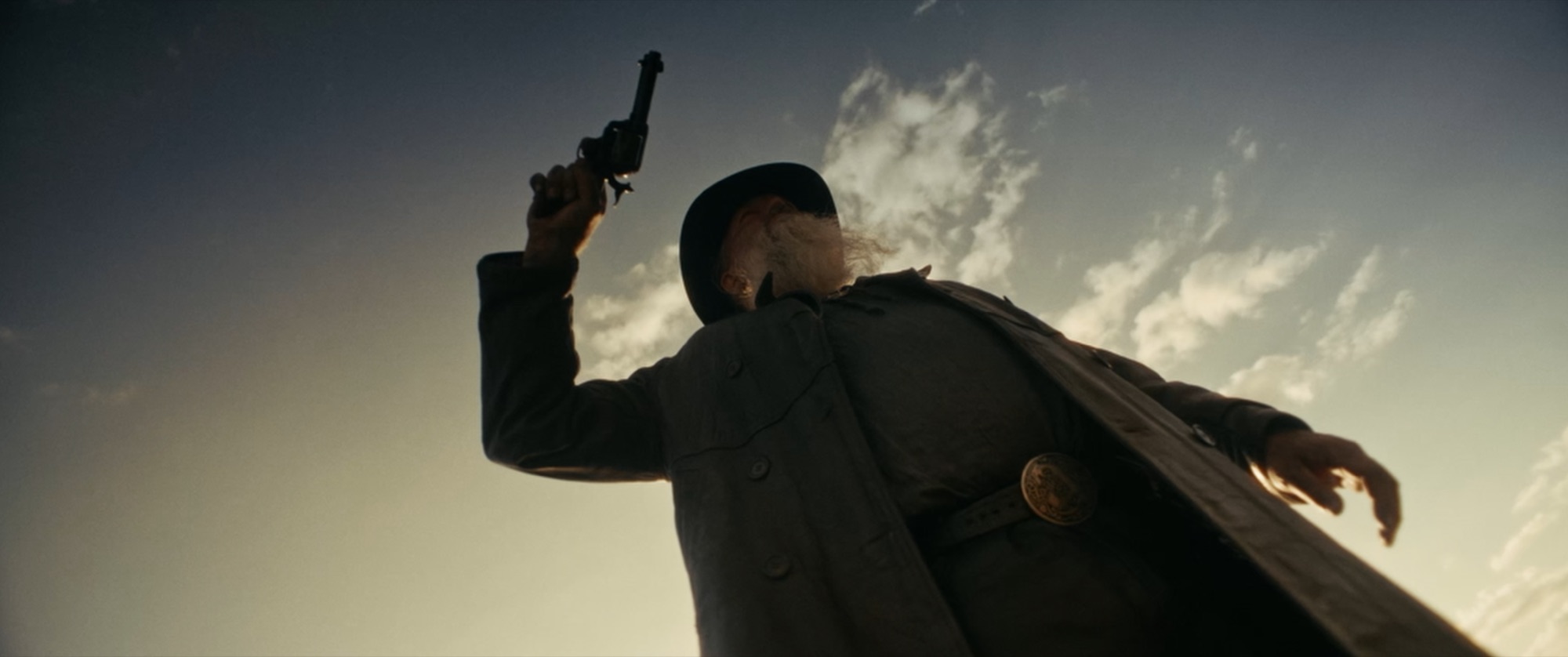 Low-angle silhouette of a long-bearded man in a duster raising a revolver to the sky - Amon Amarth